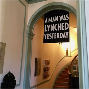 ID: Hallway with a rounded high doorway. From the doorway hangs a black flag with the words a man was lynched today in white lettering.